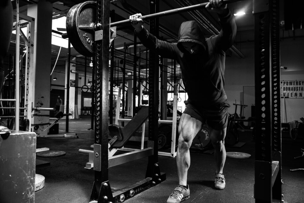 A man with his hood up stood leaning against the barbell on the squat rack mentally preparing to do a set