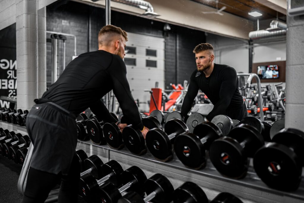 Man in gym leaning against dumbell rack looking into the mirror preparing to do his warm up sets